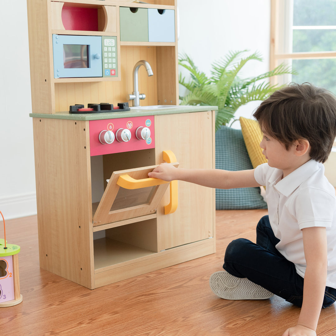 Boy sitting on the floor opening the play oven on the Teamson Kids Little Chef Florence Classic Play Kitchen with 5 Kitchen Accessory Toys, Wood Grain