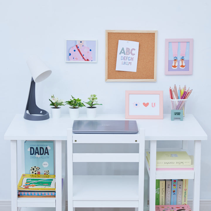 Close up of the desktop of a children's desk with a lamp, laptop, three plants and supplies against a white wall.