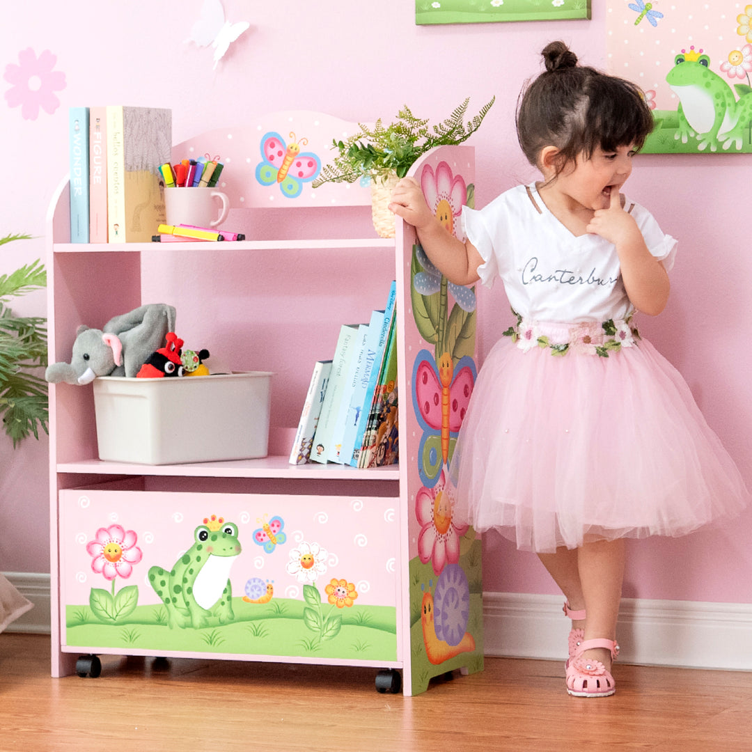 A girl in a tutu standing next to the Fantasy Fields Magic Garden Kids Wooden Toy Organizer with Rolling Storage Box, Pink bookcase.