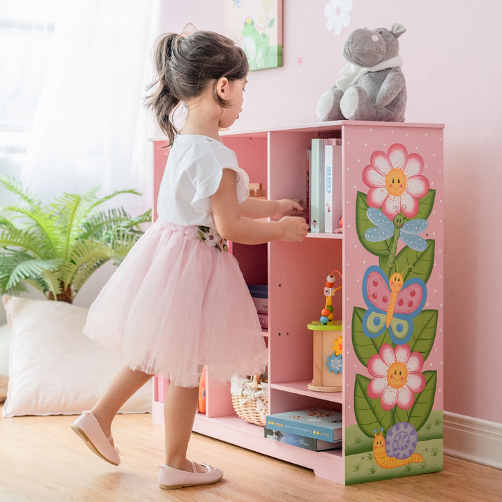 A girl in a pink dress stands beside her Fantasy Fields Kids Painted Wooden Magic Garden Adjustable Cube Bookshelf, filled with books and toys.
