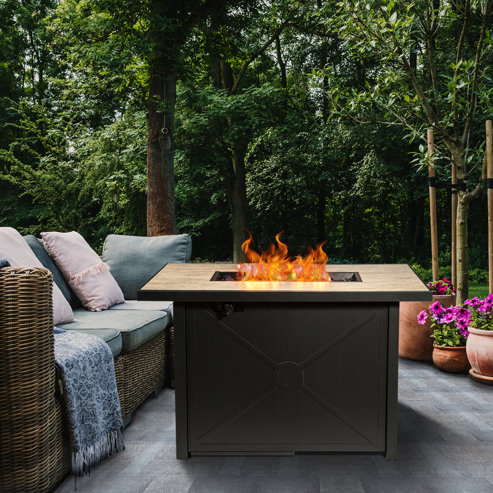 A cozy outdoor seating area with a Teamson Home 42" Outdoor Rectangular Propane Gas Fire Pit with Steel Base, Black/Stone amid lush greenery, featuring sturdy construction.