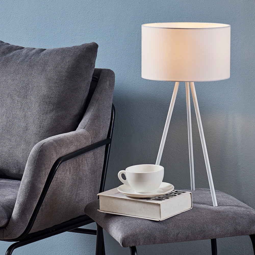 A cozy reading nook with a gray armchair next to a Teamson Home 19.7" Eli Tripod Table Lamp with Drum Shade, White, on a table