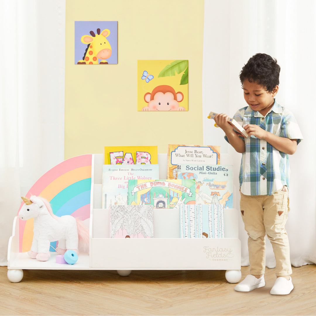 A young boy standing in front of a Fantasy Fields Rainbow Wooden Display Bookcase, White.