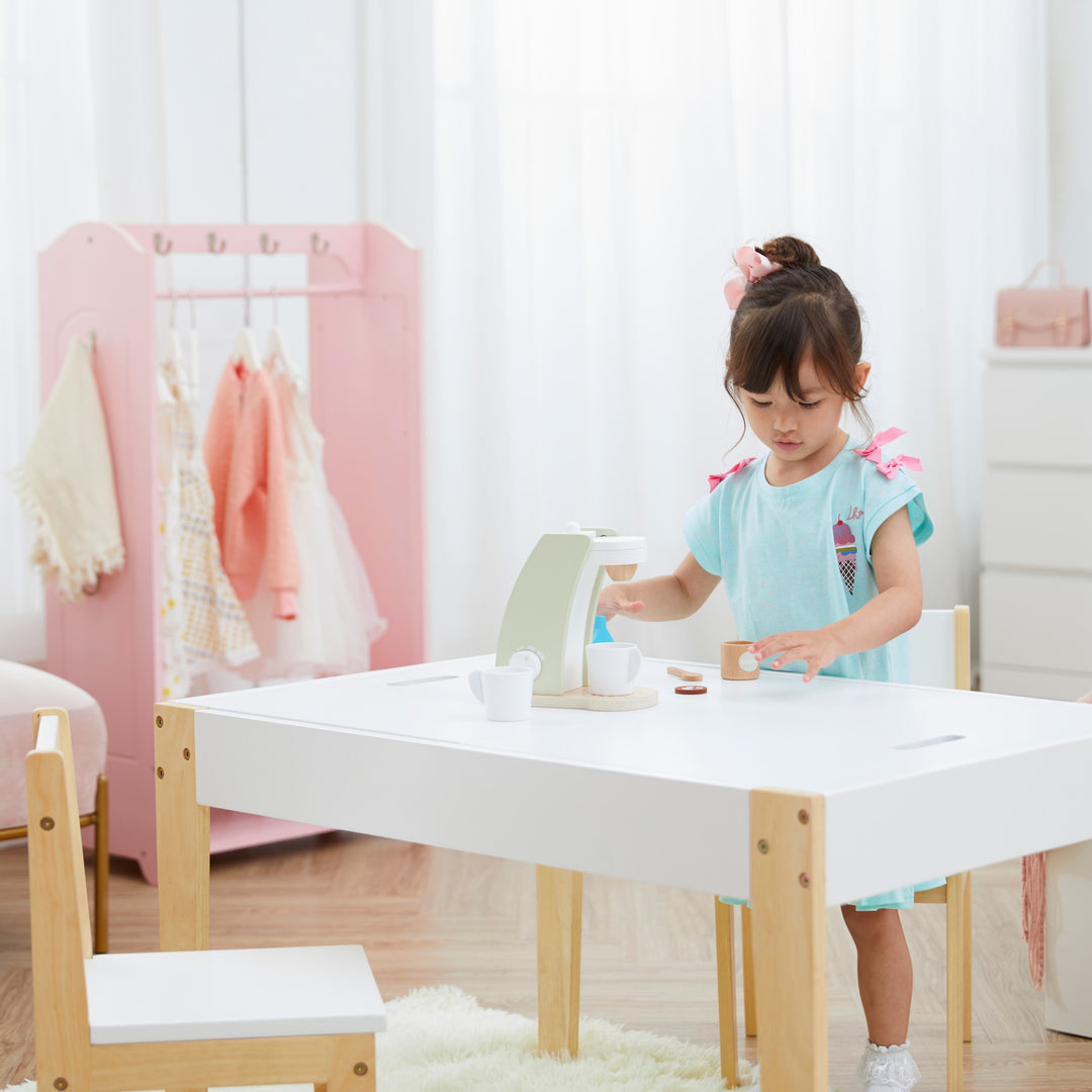 A little girl playing with her her pretend coffee maker on a white and wood child-sized table.