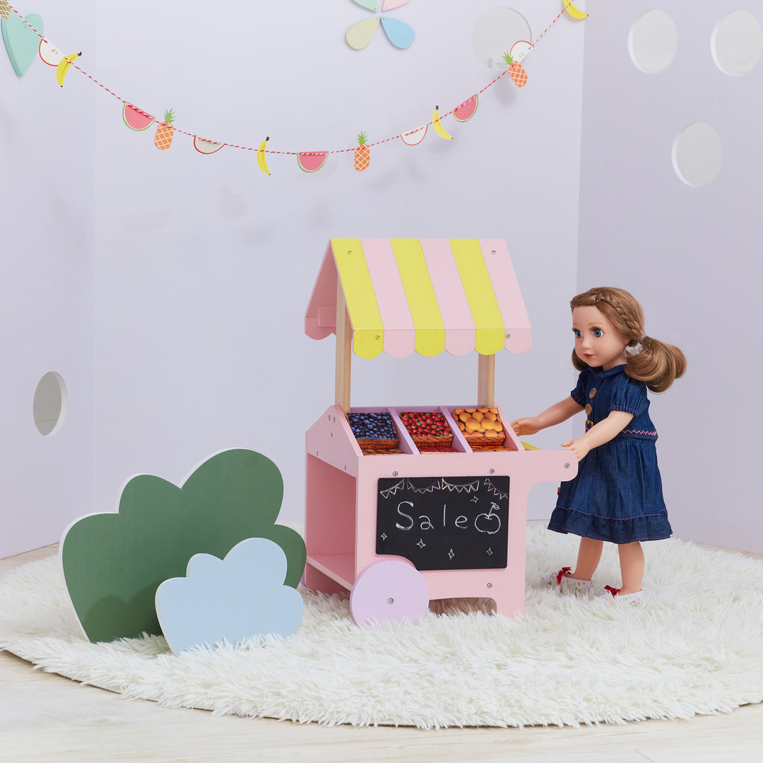 A doll in a blue dress standing at a pink and yellow fruit stand with a chalkboard and fruit boxes.
