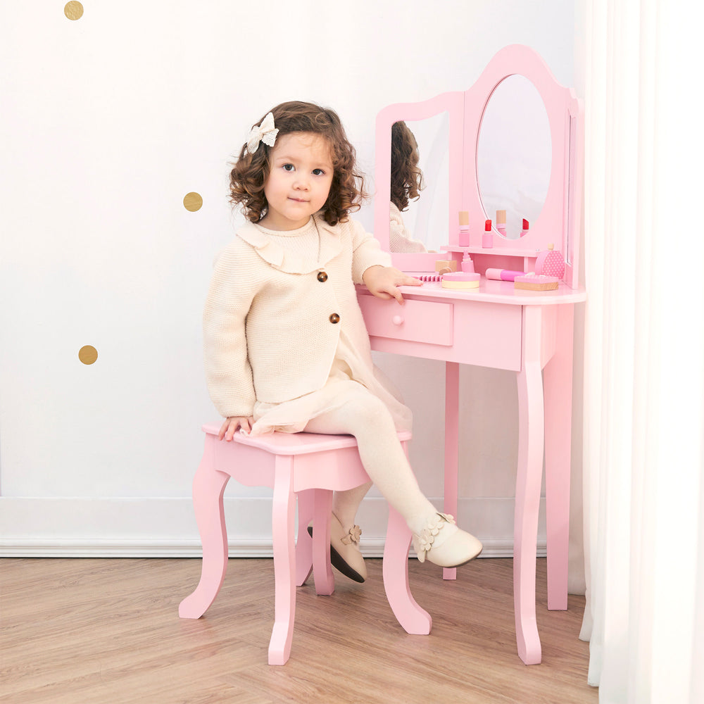 A little girl dress in ivory sitting at a pink vanity table and stool with a tri-fold mirror.