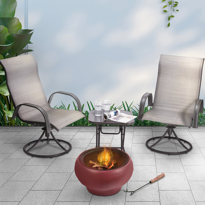 Two outdoor chairs facing a Teamson Home Outdoor 21" Wood Burning Fire Pit with Grill Grate and Faux Concrete Base, Maroon, against a backdrop of plants and a cloudy sky.
