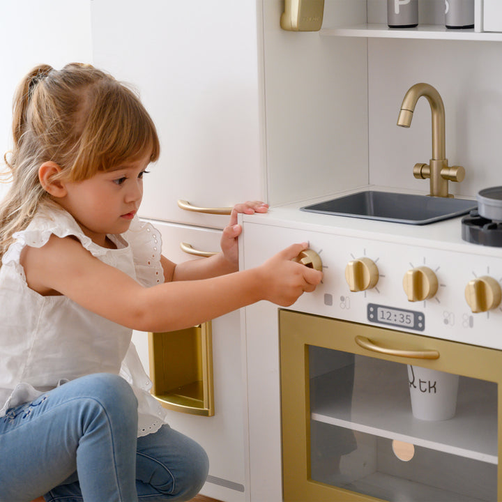 A young girl plays with a Teamson Kids Little Chef Mayfair Classic Kids Kitchen Playset with 11 Accessories, White/Gold, pretending to turn on the play oven.