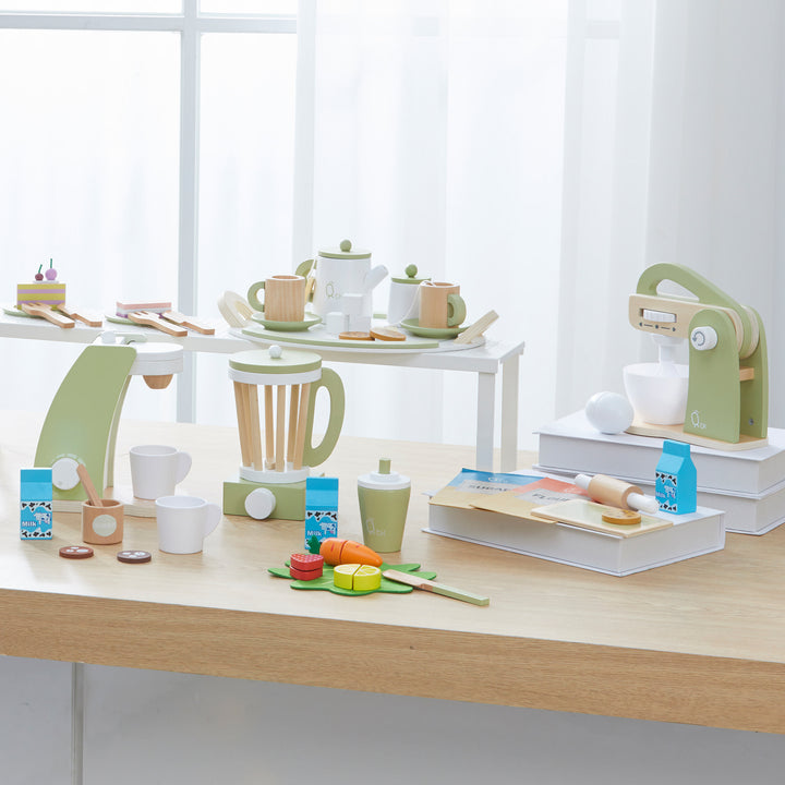 Kitchenware, pretend ingredients, and Teamson Kids Little Chef Frankfurt Wooden Blender Play Kitchen Accessories displayed on a wooden table and white shelves in a bright room.
