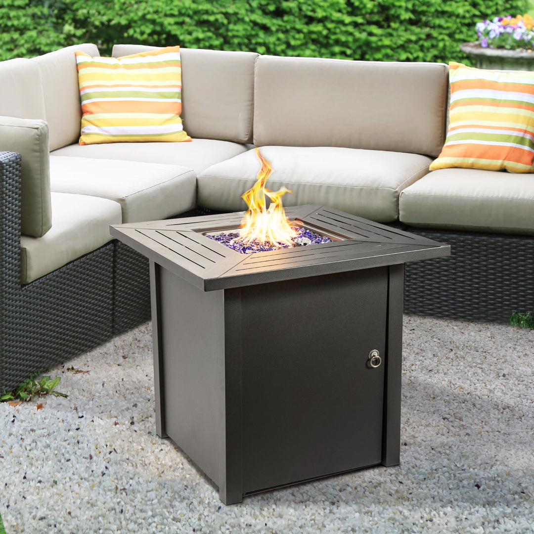 A Teamson Home Outdoor Square 30" Propane Gas Fire Pit with Steel Base, with a flame in the center, placed on a gravel patio area next to an outdoor sectional sofa with colorful pillows.