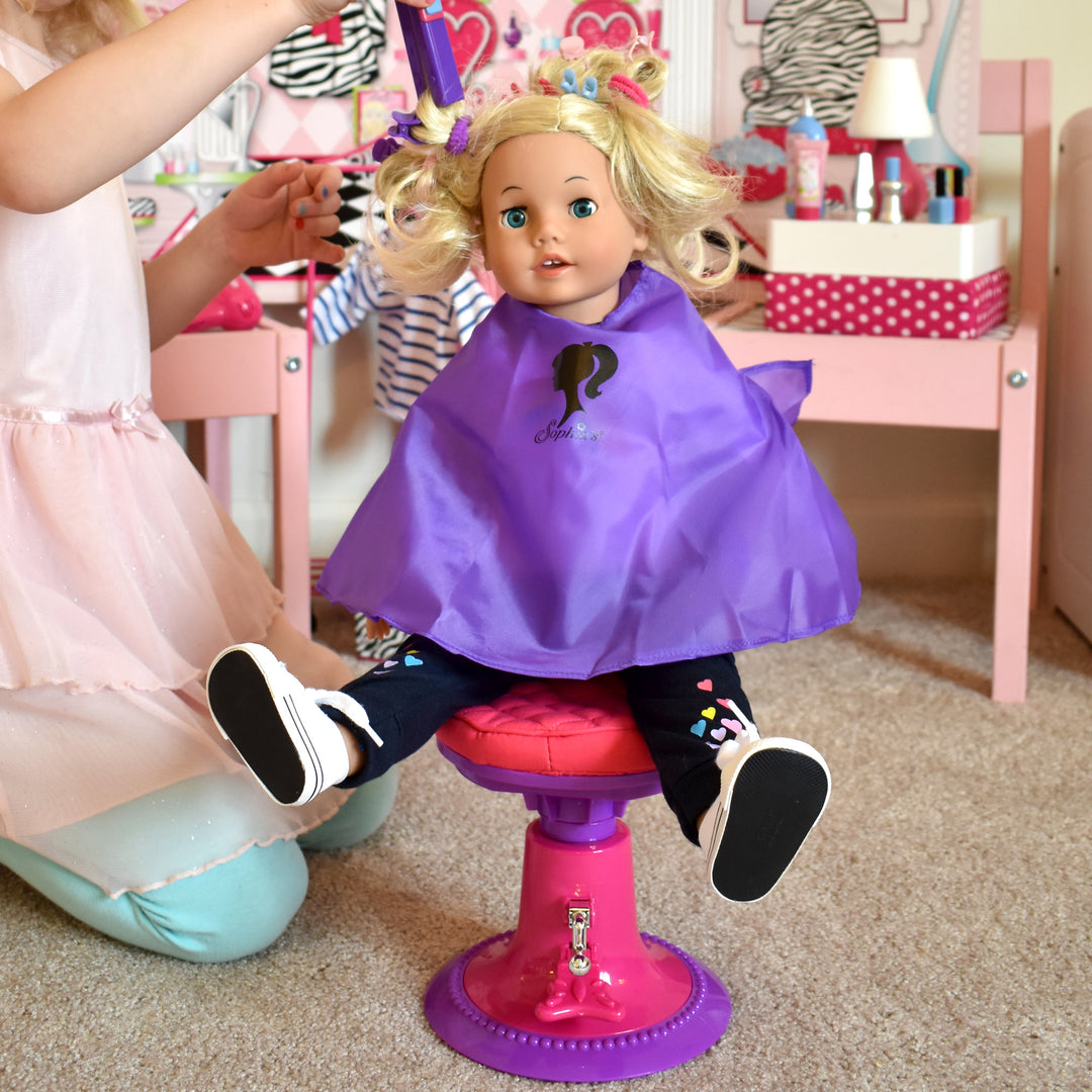 A little girl kneeling while she does her 18" blonde doll's hair. The doll is sitting in a salon chair and wearing a purple smock.