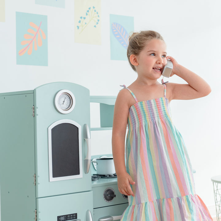 A young girl in a striped dress plays with a Teamson Kids Little Chef Westchester Retro Kids Kitchen Playset, Mint next to a play kitchen set.