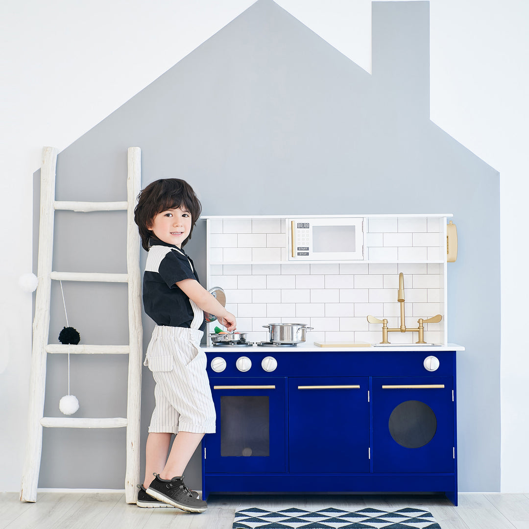 A child pretends to be a little chef standing by a Teamson Kids Little Chef Berlin Play Kitchen with Cookware Accessories, White/Blue designed to look like a miniature version of a modern kitchen.