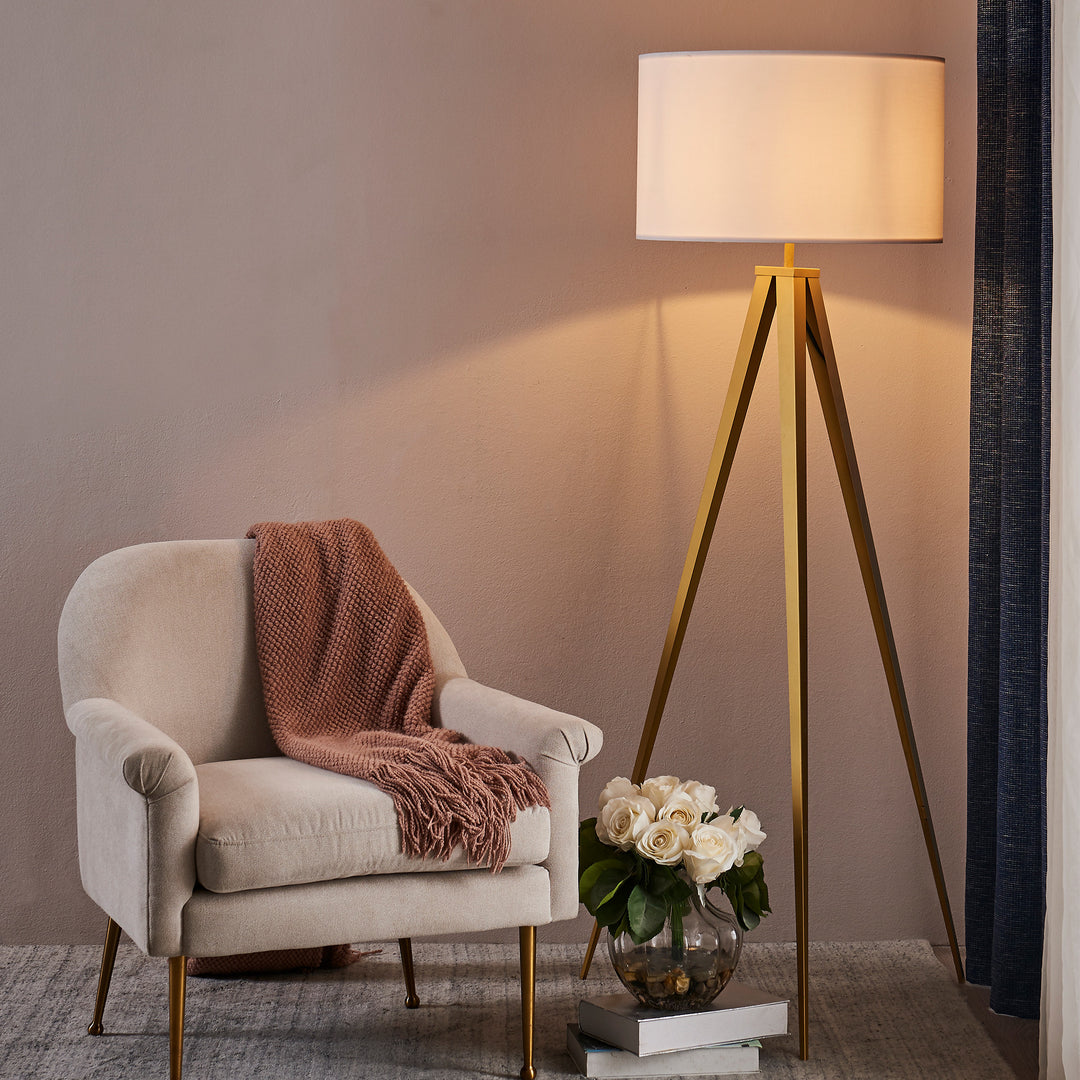 Elegant interior setup with a comfortable armchair, a knitted throw, the Teamson Home Romanza 62" Postmodern Tripod Floor Lamp with Drum Shade, Matte Gold/White, and a vase of white roses.