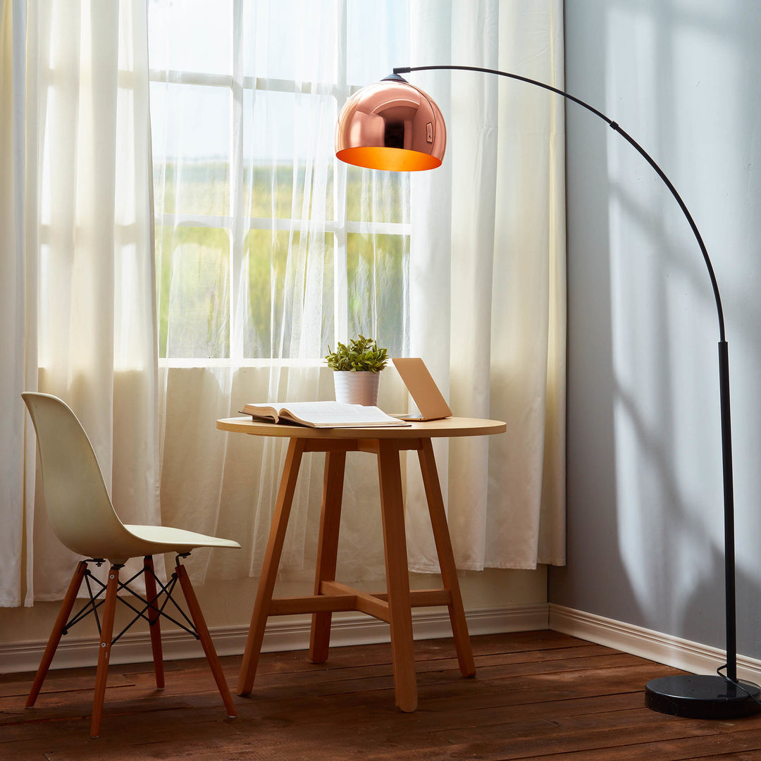 A Teamson Home Arquer Arc 66" Metal Floor Lamp with a rose gold shade and a wooden table.