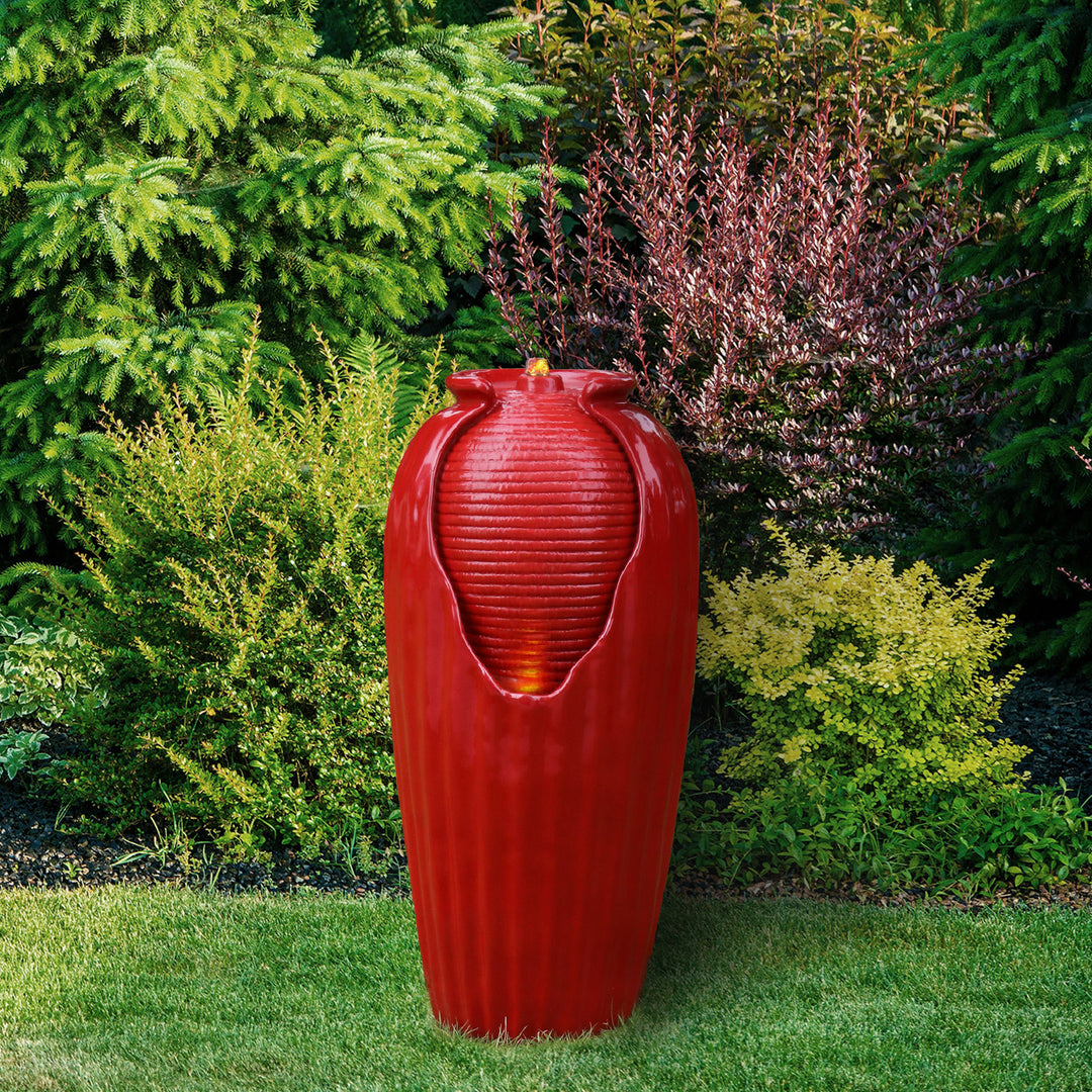 A Teamson Home Indoor/Outdoor Contemporary Glazed Contoured Vase Water Fountain with LED Lights, Red stands in front of various shrubs in a garden, serving as a striking outdoor accent.