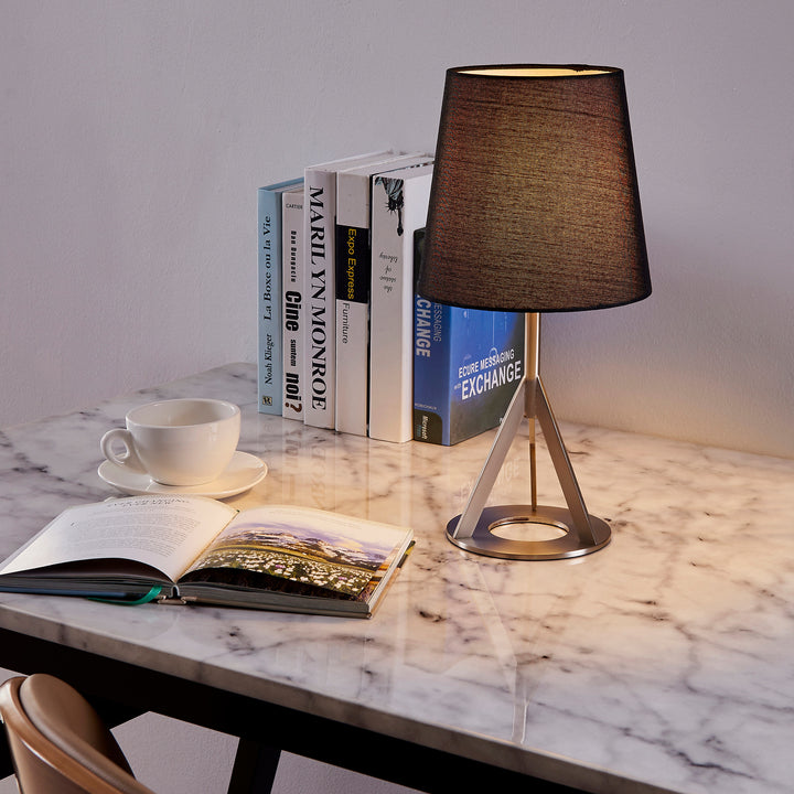A Teamson Home's Aria 15" table lamp with a black linen-like shade and a geometric base in nickel on a faux white marble tabletop.
