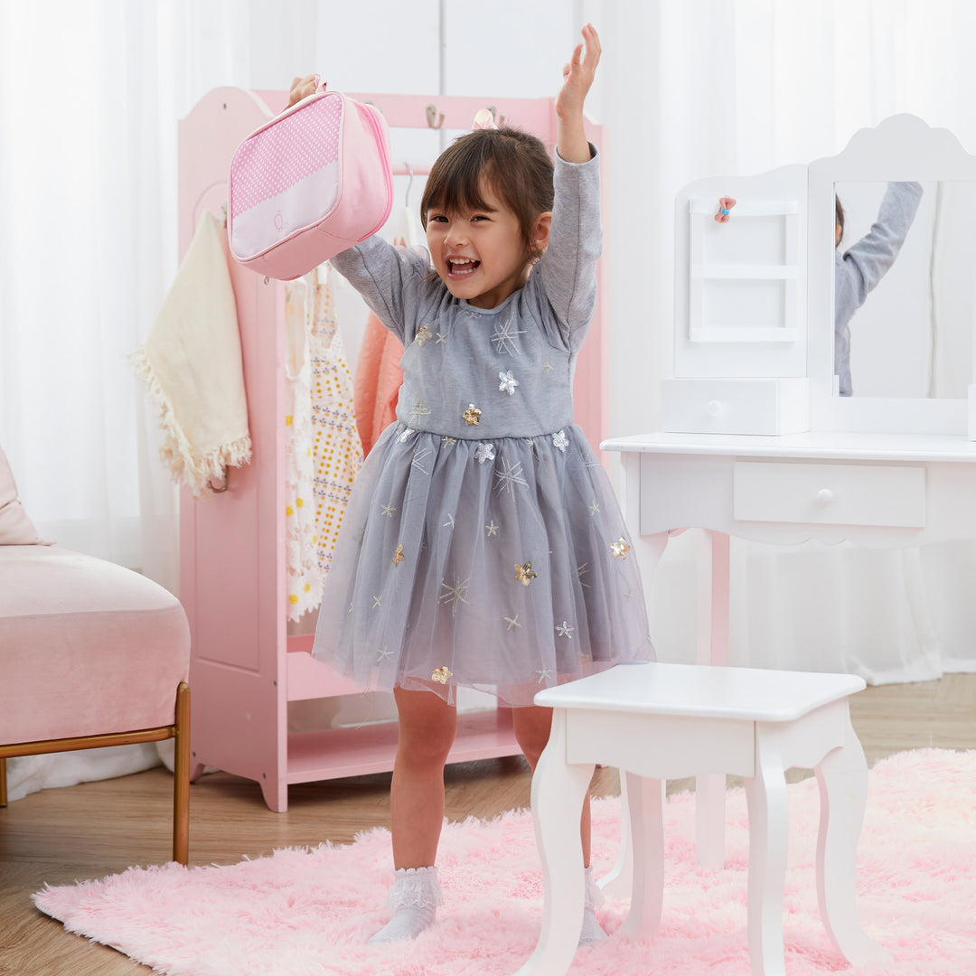 A happy little girl holding the pink make-up bag up in the air while she stands in front of a pink wardrobe and next to a white vanity table and stool.
