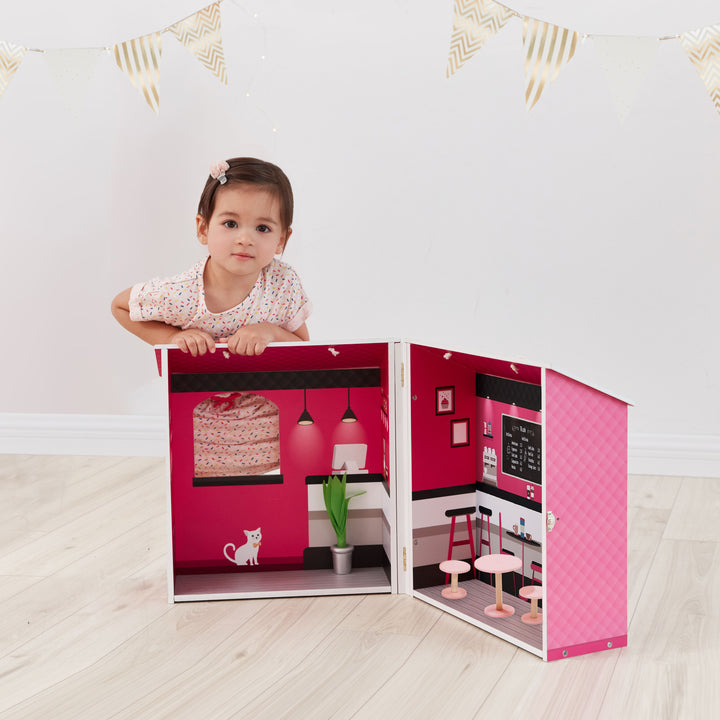 A little girl sitting behind of Olivia's Little World Dreamland City Café Dollhouse, Pink with accessories.