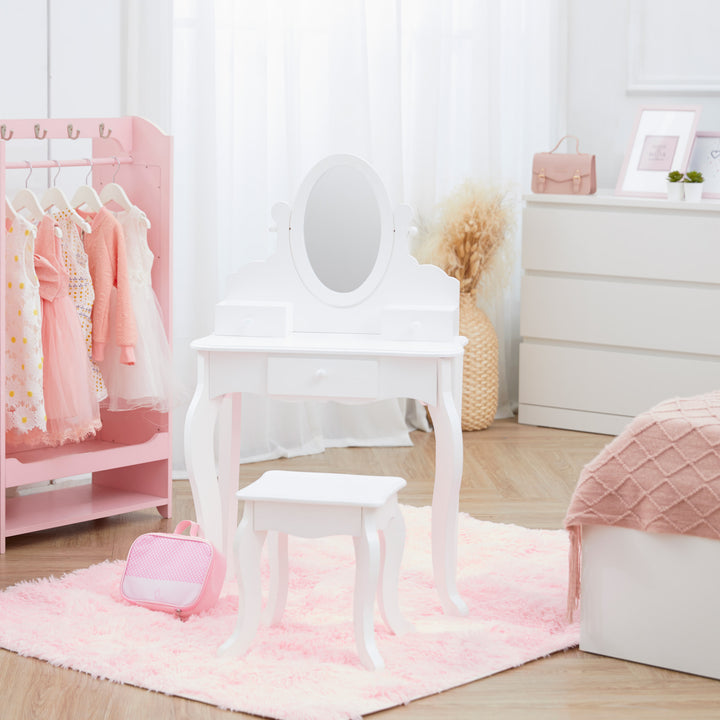 A white vanity table with mirror and matching stool on a pink rug in a bedroom with a pink wardrobe with dresses hanging on the left and a bed with a pink blanket on the right.
