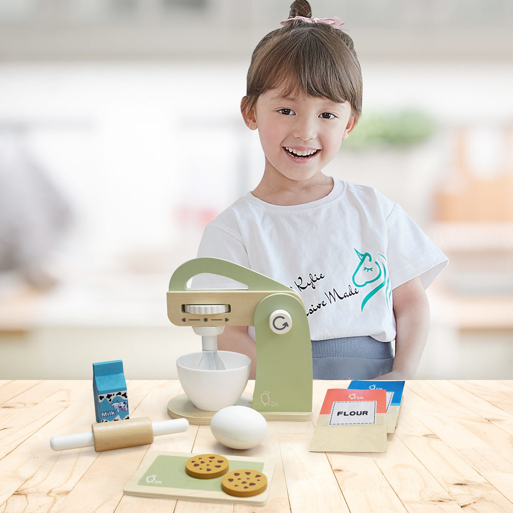 A smiling child playing with a Teamson Kids Little Chef Frankfurt Wooden Mixer Play Kitchen Accessories set and pretend ingredients on a wooden table.