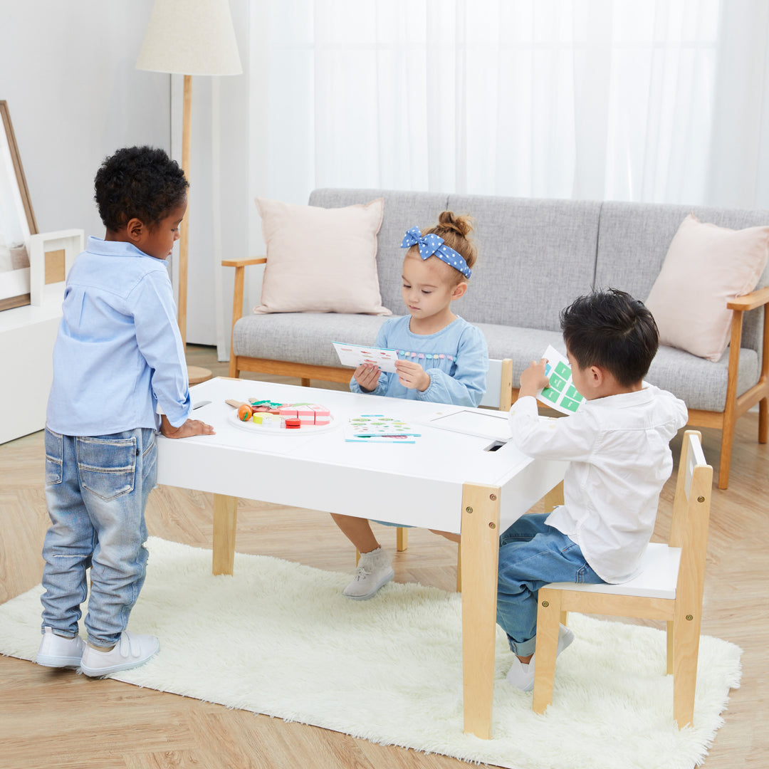 A little boy standing, a little boy sitting and a little girl sitting around a child-sized white and wooden table in a playroom.