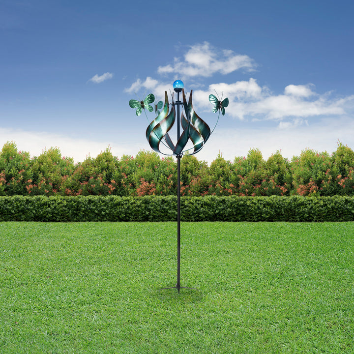 A colorful, Teamson Home Outdoor Solar Tulip and Butterfly Kinetic Windmill Sculpture with LED Light, Teal installed in a green lawn against a backdrop of hedges and a clear blue sky.