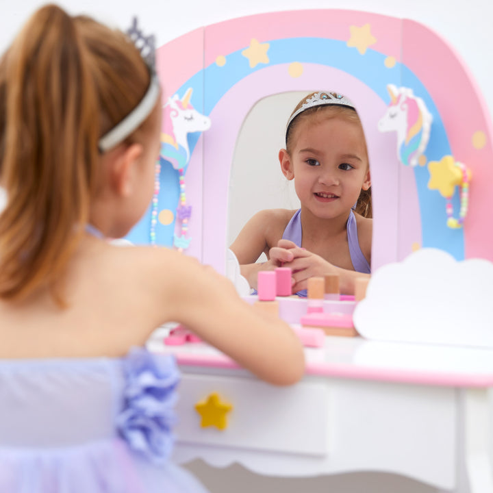 A little girl sitting and looking into the mirror of her rainbow and white vanity.