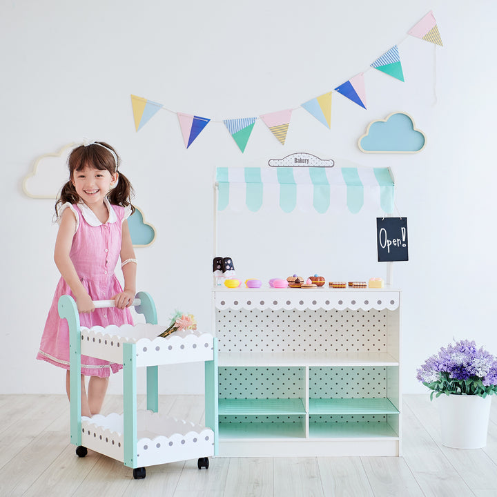 A young girl stands next to her Dream Bakery Shop Stand filled with treats, and pushing a cart with flowers