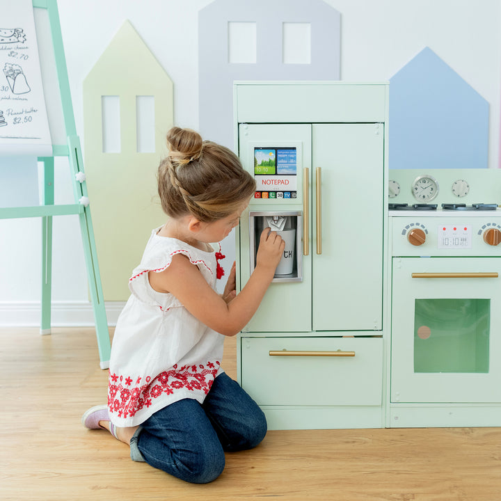 A young girl is engaged in play with a Teamson Kids Little Chef Charlotte Modern Play Kitchen, Mint/Gold toy atm machine, featuring realistic details and interactive features, integrated into a wooden kids play kitchen set, depicting a scene of imaginative domestic and financial role-playing.