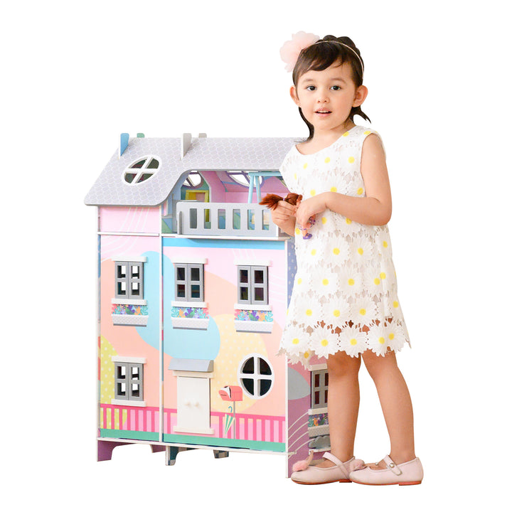 A little girl standing in front of a Teamson Kids Sunroom Dollhouse with 11 Accessories, Multicolor.