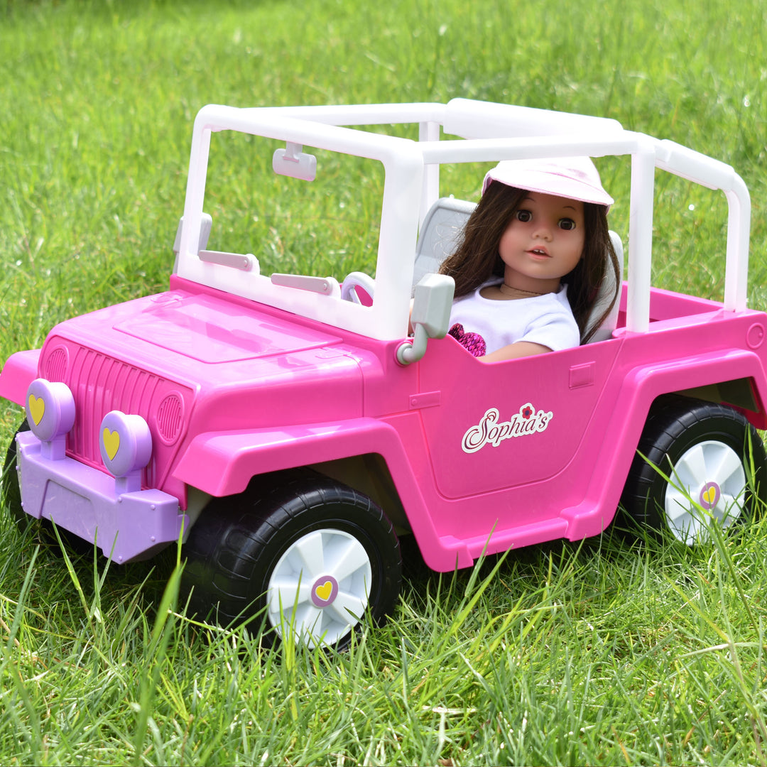 A brunette 18" doll sitting in the driver's seat  of a pink 4x4 truck in the grass.