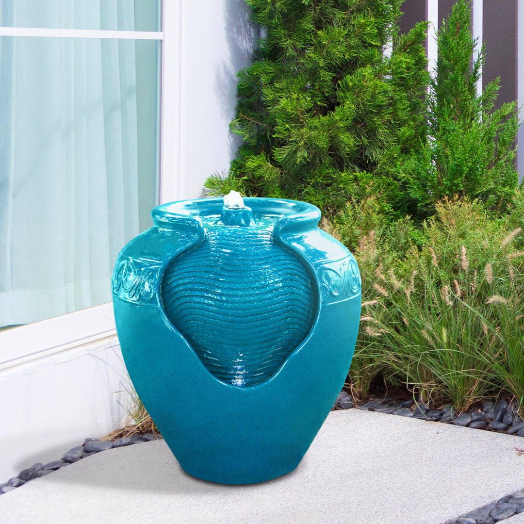 A teal Teamson Home Outdoor Glazed Pot Floor Fountain with LED lights, in the shape of a vase on a patio beside green shrubbery.