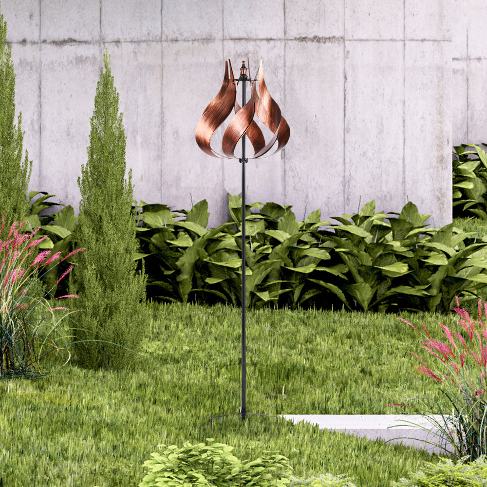 A Teamson Home Outdoor Tulip Kinetic Windmill Sculpture, Tangerine with a twisted design, displayed in a lush garden setting against a concrete wall.