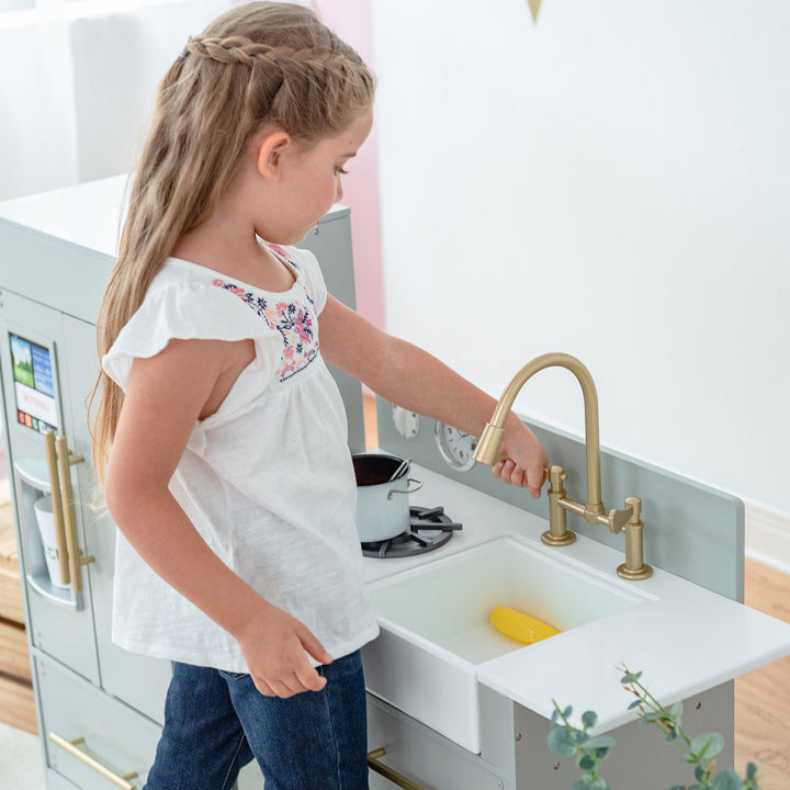 A young girl playing with a Teamson Kids Little Chef Charlotte Modern Play Kitchen, Silver Gray/Gold.