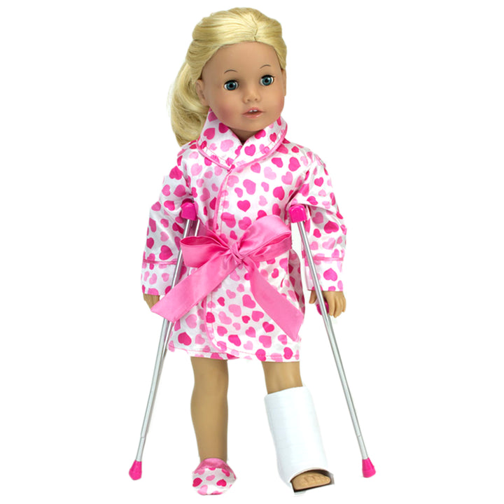 A blonde 18" doll with a pink hearts robe with a cast on her left leg standing on crutches.