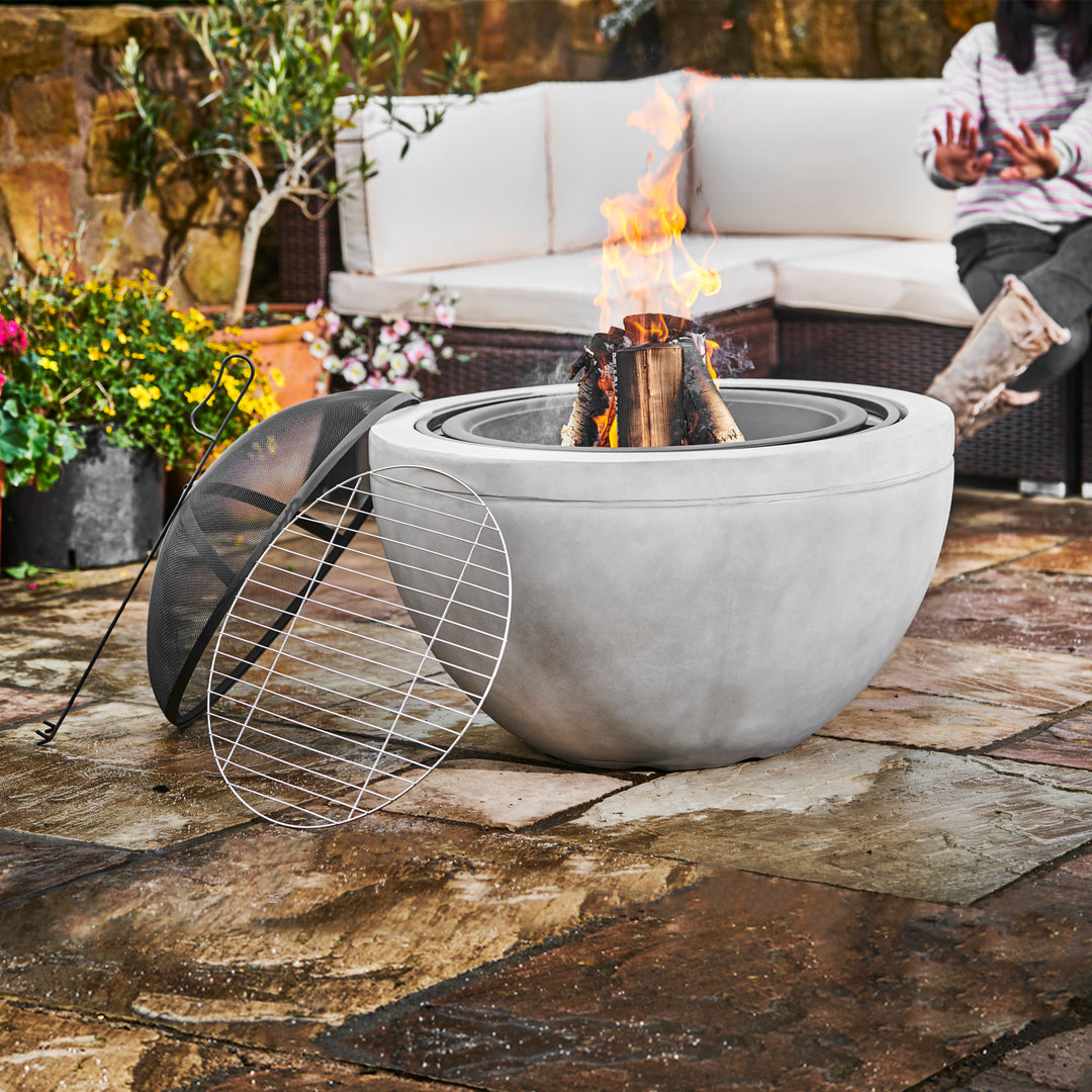 Teamson Home 30" Outdoor Round Wood Burning Fire Pit with Faux Concrete Base, Gray with a metal screen beside it, set on a patio with a person lounging in the background, enhancing the outdoor decor.