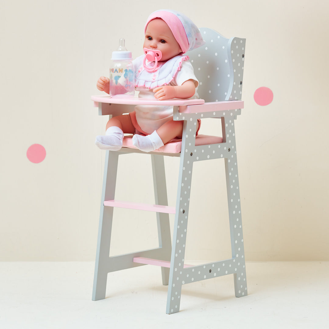 A baby doll sitting in a pink and grey with white polka dots baby doll high chair with a bottle on the tray.