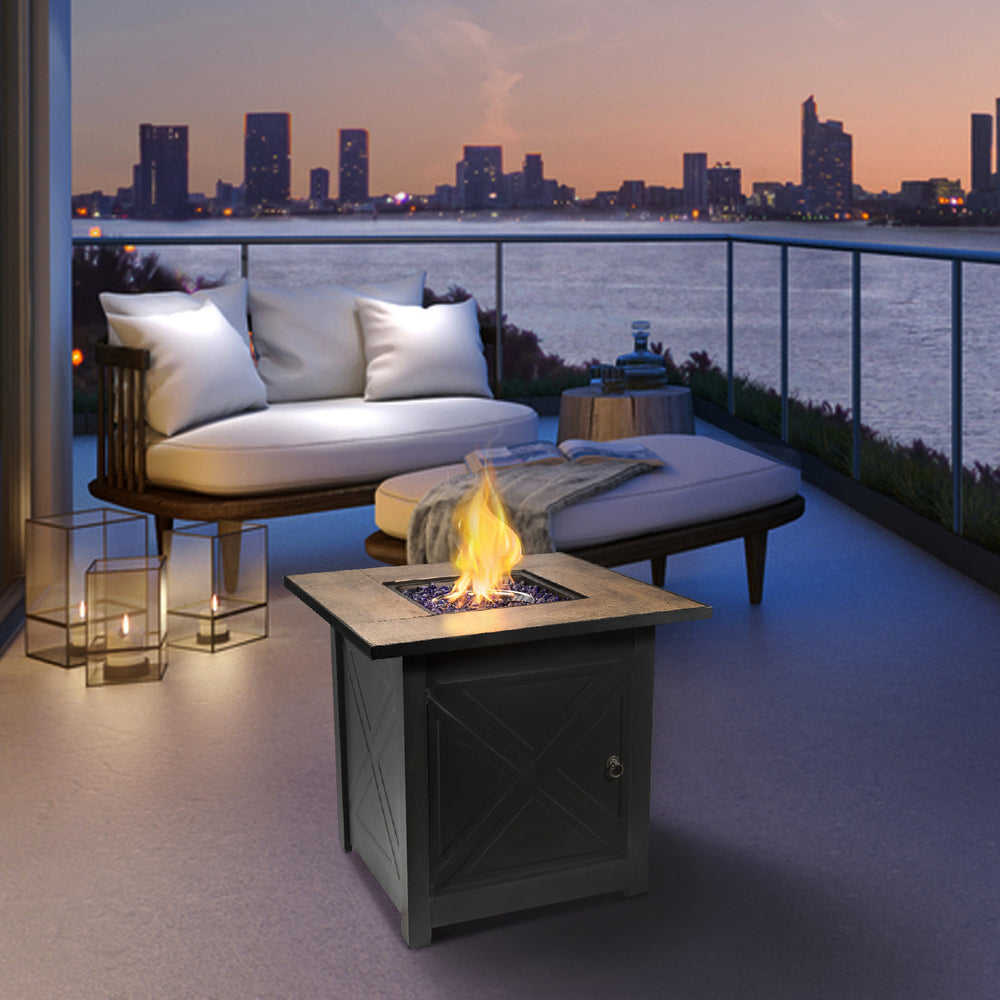 Teamson Home Outdoor Square 30" Propane Gas Fire Pit with Steel Base, Black table and lounge seating overlooking a city skyline at dusk.