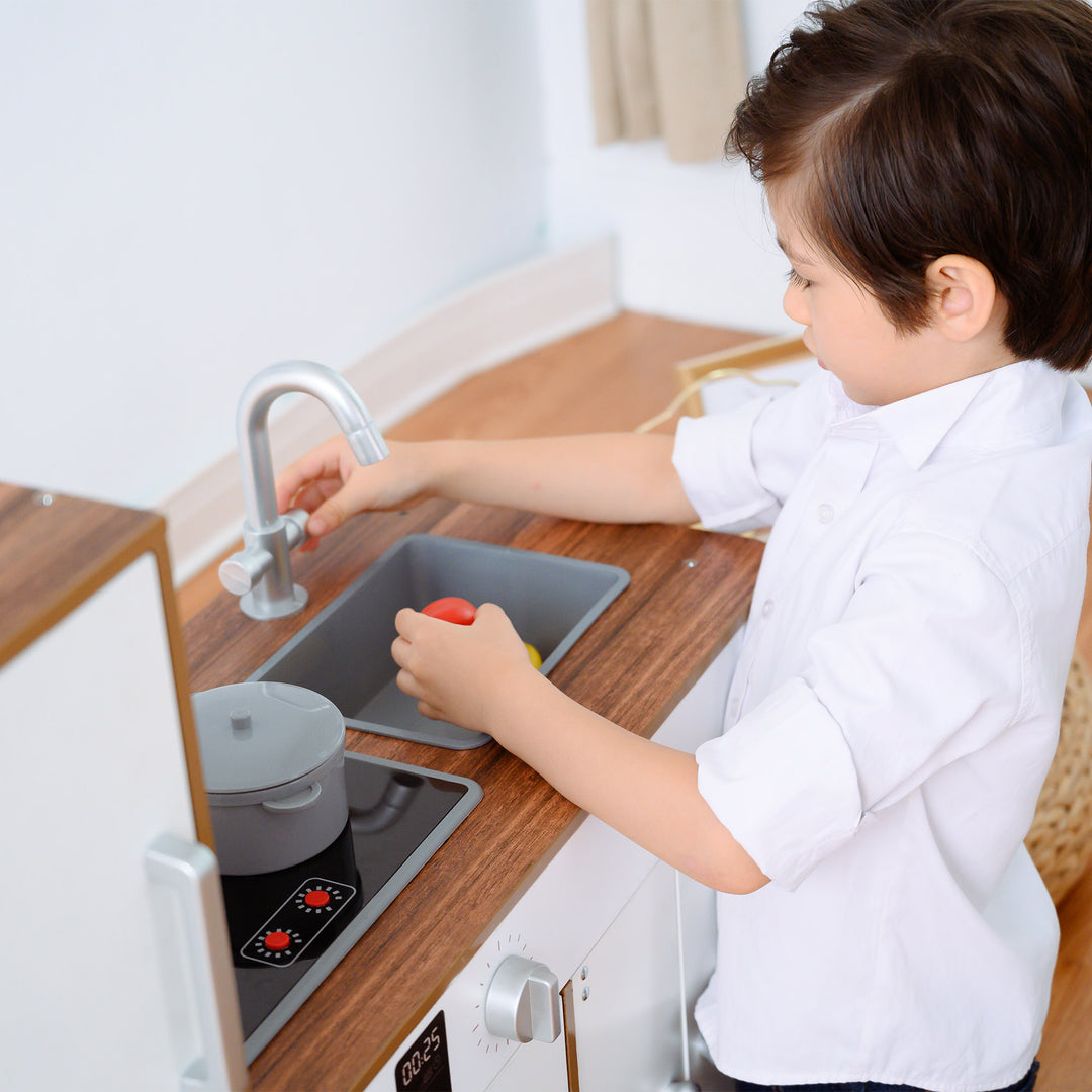A child pretending to wash dishes at the Teamson Kids Little Chef Palm Springs Classic Kids Play Kitchen with Accessories, Natural/White designed with kids-sized dimensions.