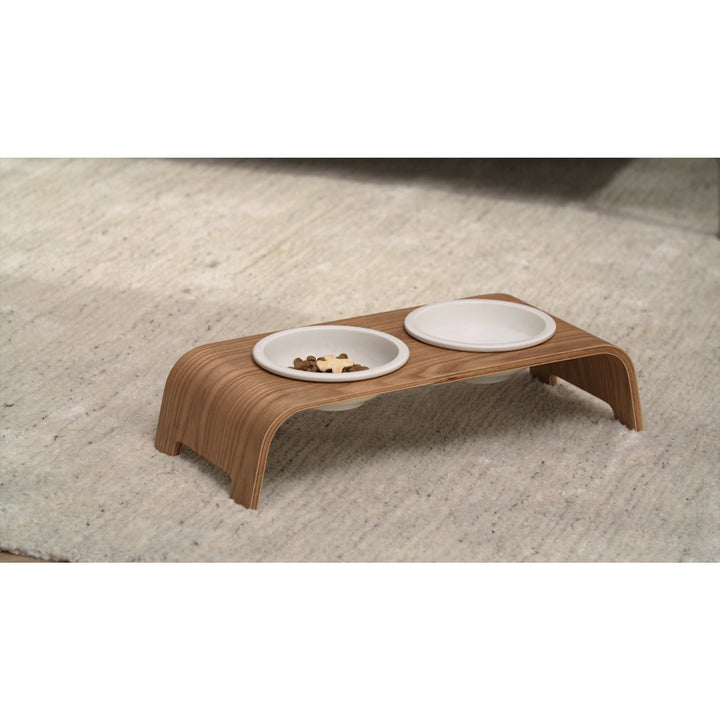 A Teamson Pets Billie Small Elevated Ash Wood Pet Feeder with two white spill-proof bowls, one containing food, on a carpeted floor.