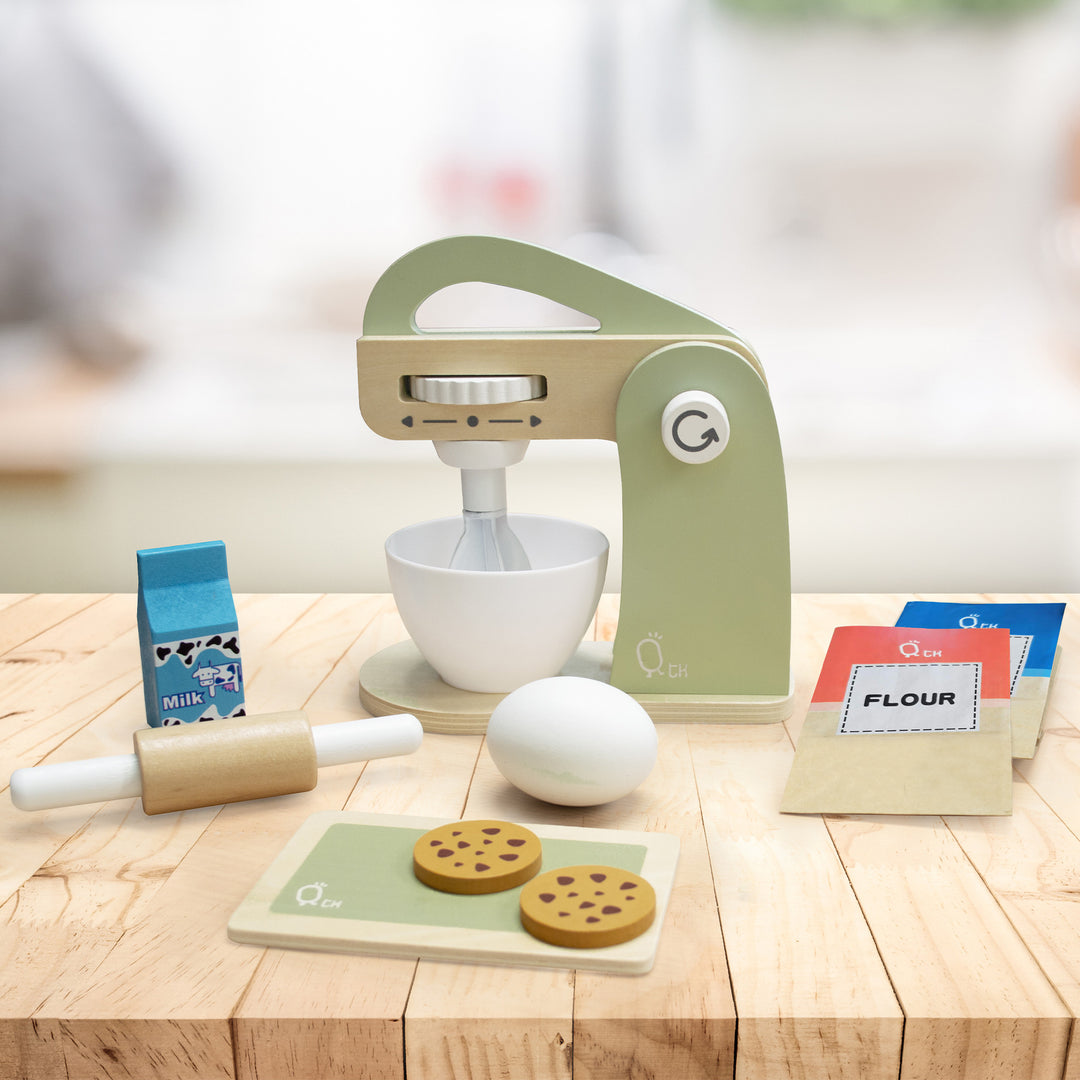 A Teamson Kids Little Chef Frankfurt Wooden Mixer Play Kitchen set with pretend ingredients including milk, flour, an egg, and cookies on a wooden tabletop.