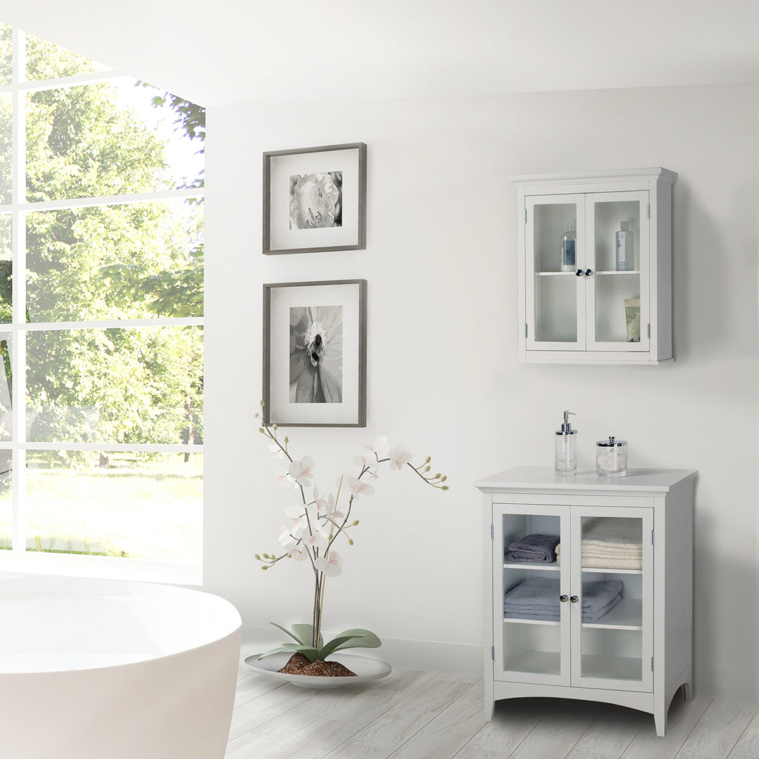 Bright bathroom interior with a bathtub, a Teamson Home White Madison Removable Wall Cabinet with 2 Doors, with framed pictures, accented by a small plant.