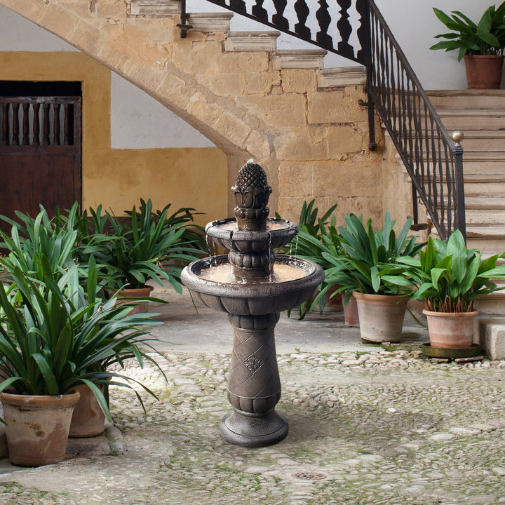 Ornate Teamson Home Deluxe Pineapple 2-Tier Waterfall Fountain in a serene courtyard surrounded by potted plants.