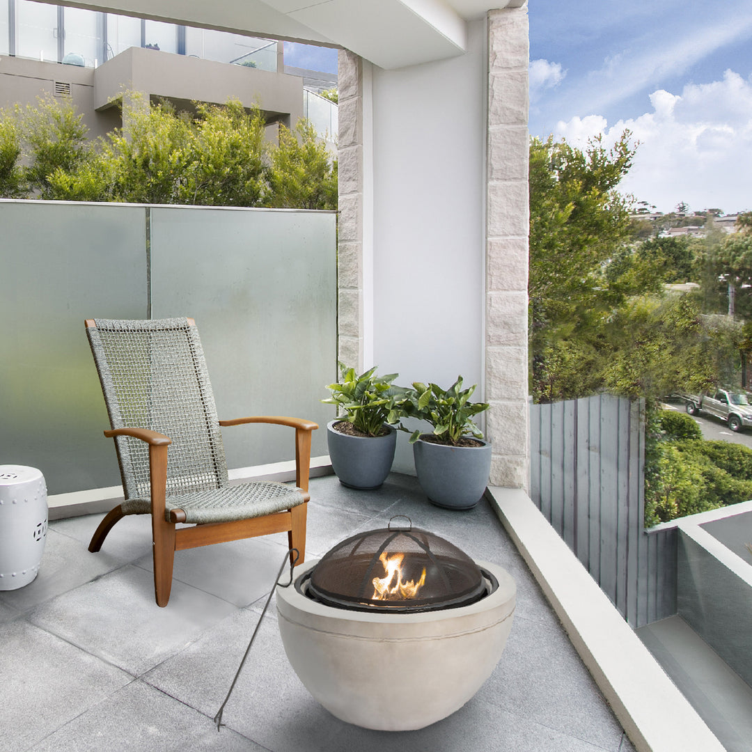 Modern balcony with a Teamson Home 30" Outdoor Round Wood Burning Fire Pit with Faux Concrete Base, Gray, single chair, and potted plants, overlooking an urban area, enhanced by outdoor decor.