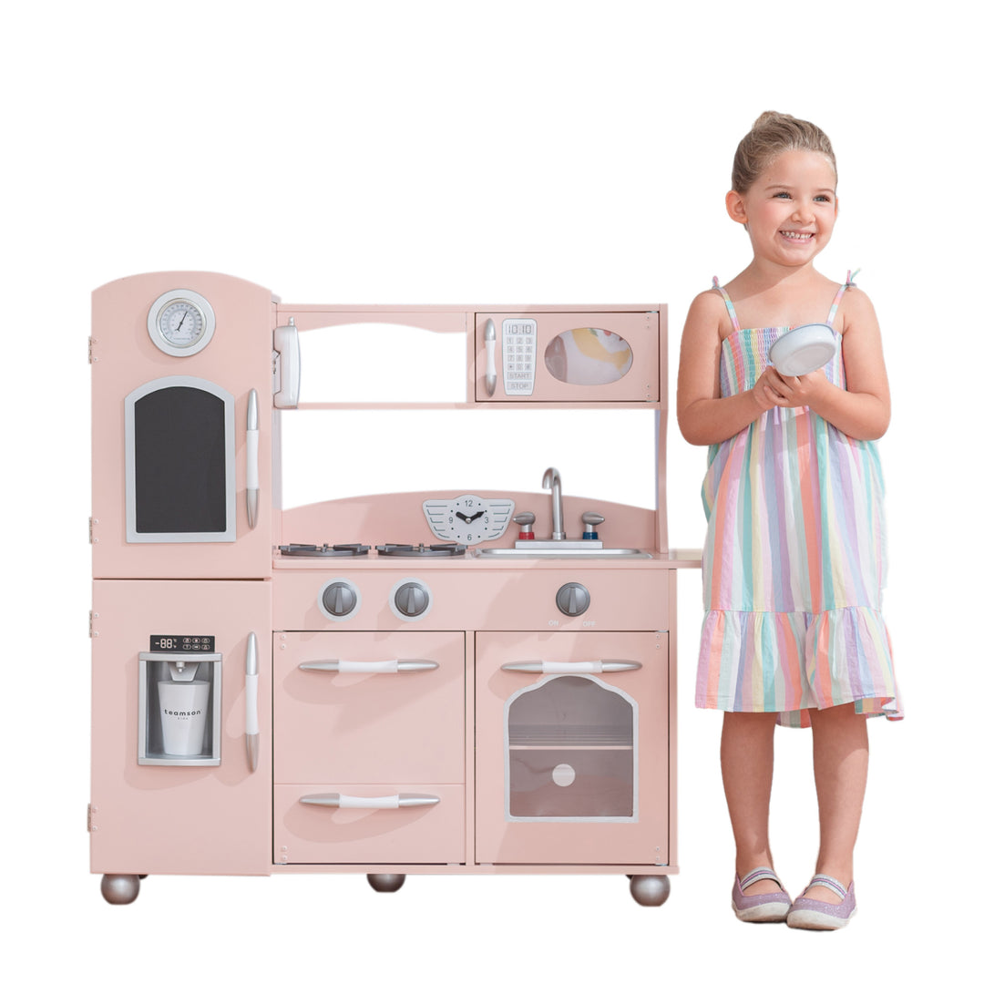 Young girl smiling beside a Teamson Kids Little Chef Westchester Retro Play Kitchen, Pink, holding a plate.