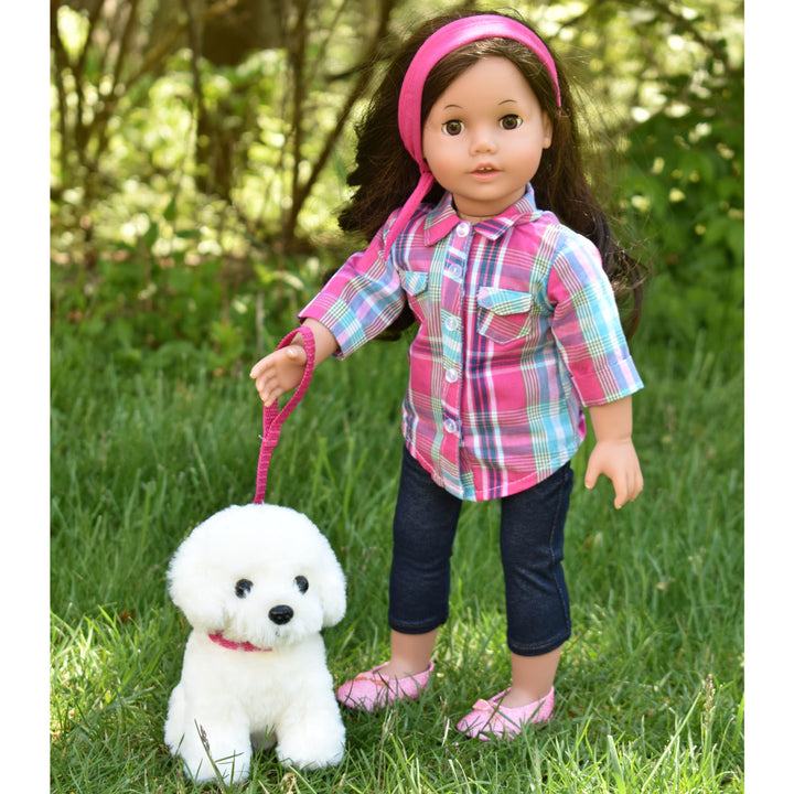 An 18" brunette doll in a plaid shirt with denim capri pants holding a pink leash with a white puppy on the end
