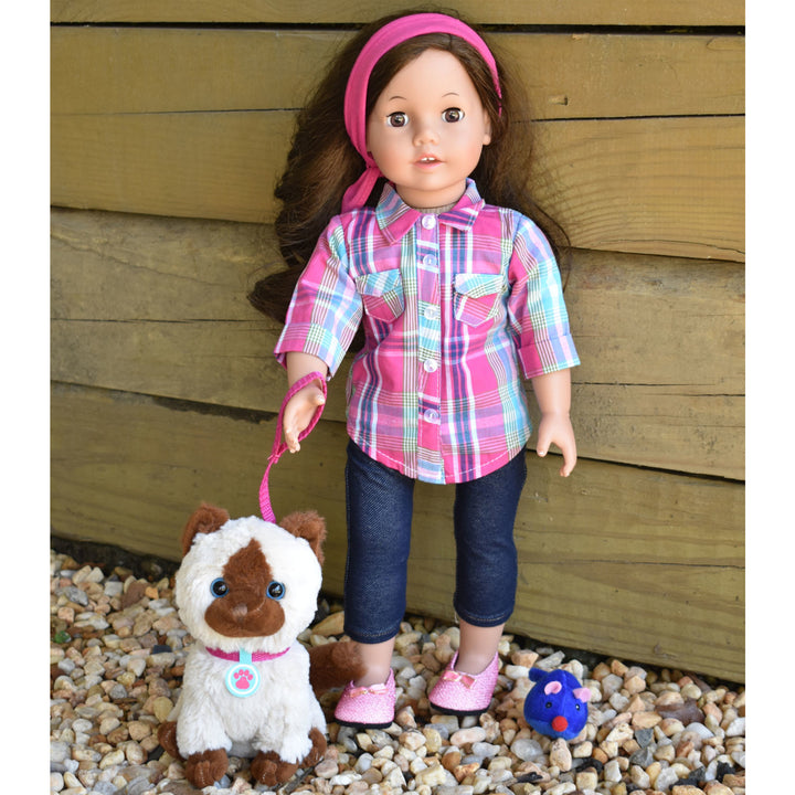 An 18" brunette doll with a plaid shirt and denim capri pants and pink shoes. She is standing on some gravel with a siamese kitten on a pink leash.
