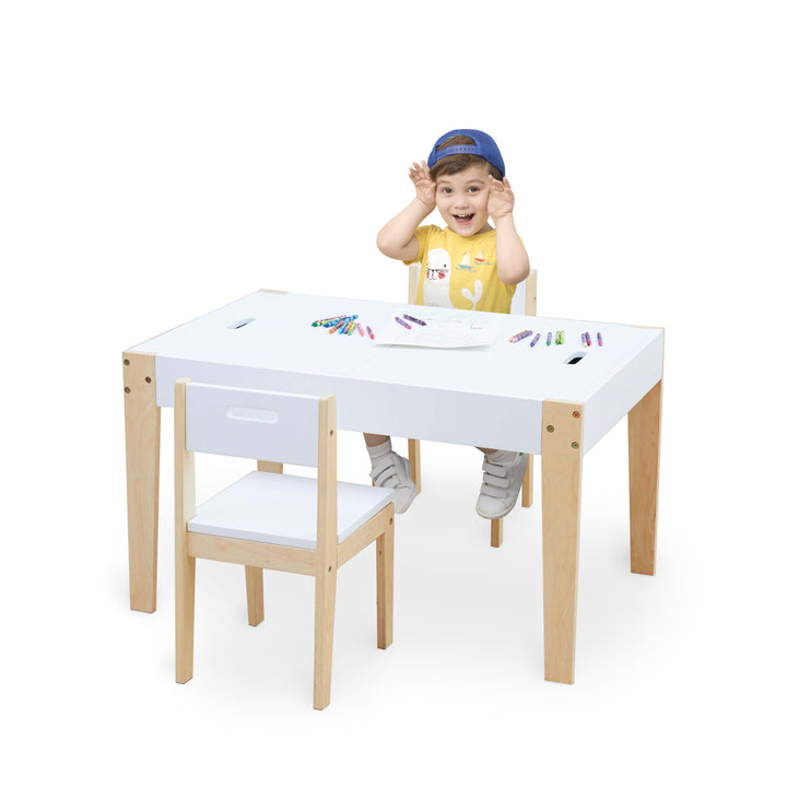 A young boy sitting at a white Fantasy Fields 3 Piece Play Table and Chairs Set with Storage and 2-Way Chalkboard Table Top, White table with crayons.