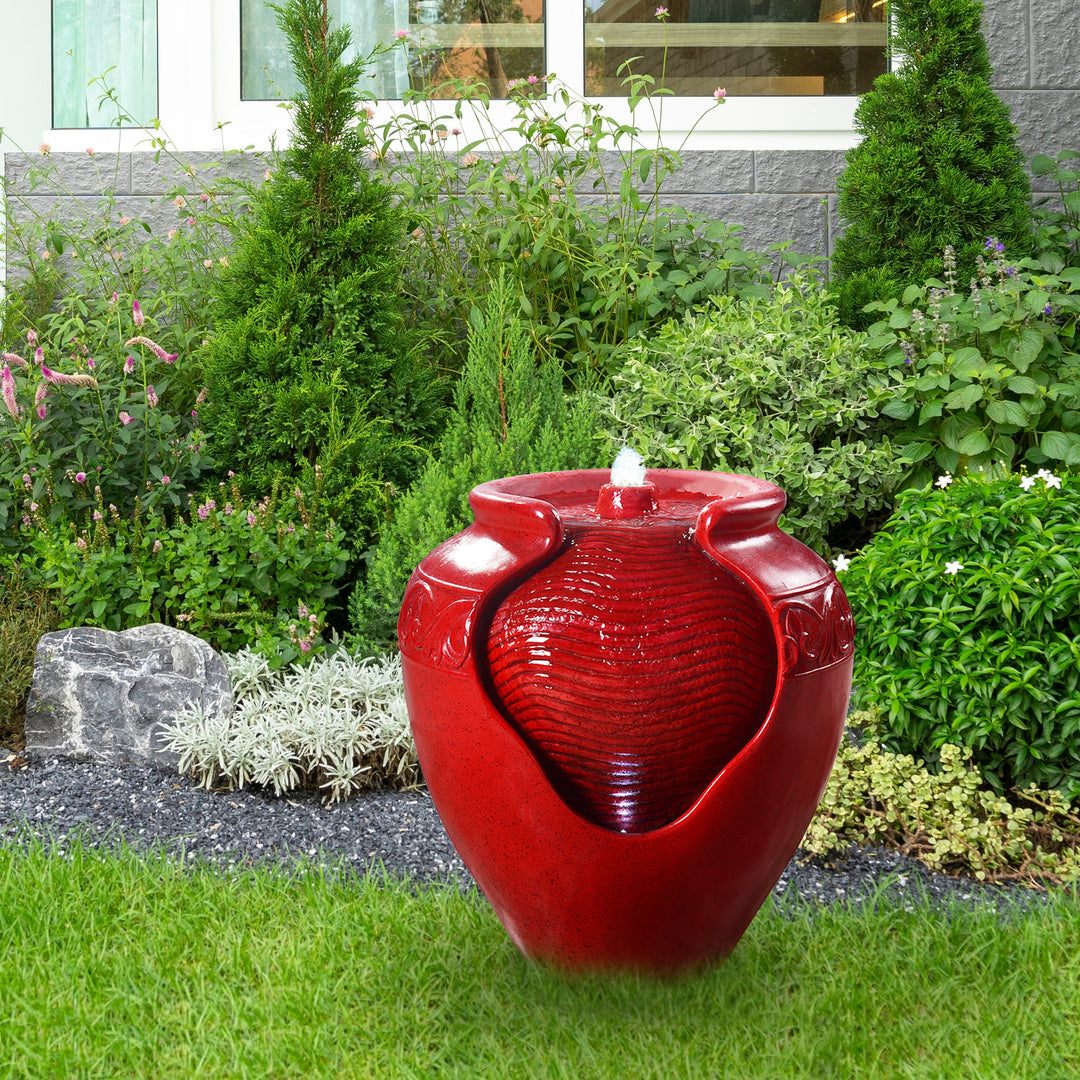 A Teamson Home Outdoor Glazed Pot Floor Fountain with LED Lights, Red in a landscaped garden.
